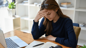 A young woman with long brown hair, wearing glasses perched on her head and a dark blue long-sleeved shirt, rests her forehead on her hand while sitting at a wooden desk. A laptop is open to her left, and a notebook with a pen and headphones sit in front of her. She appears to be looking down or has her eyes closed, suggesting contemplation or perhaps fatigue. The background includes a white shelving unit with various items and a potted plant.