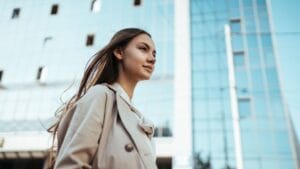 A poised woman with flowing brown hair stands against a backdrop of sleek glass skyscrapers, her beige trench coat neatly buttoned as she gazes thoughtfully into the distance, embodying confidence amidst the crisp, sunlit urban scenery.