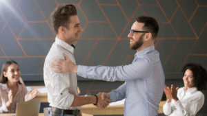 The image shows two men in an office setting, one is congratulating the other with a handshake and a pat on the back. Two women are visible in the background, smiling and applauding. This suggests a positive and supportive workplace environment.