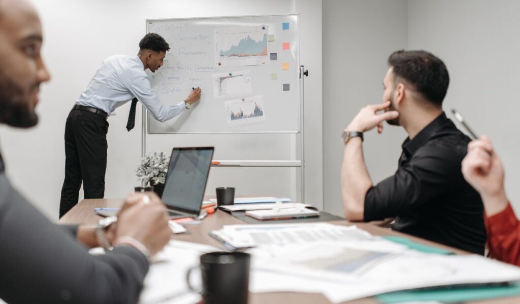 A professional meeting is taking place in a modern office setting. A man in a light blue shirt and tie is standing at a whiteboard, writing notes and pointing to various graphs and charts related to "COVID-19 Vaccination." The whiteboard contains data visualizations, including bar graphs and line charts, indicating an analysis of vaccination trends or related topics. Several team members are seated around a conference table, attentively observing the presentation. The table is equipped with laptops, documents, and coffee mugs, suggesting a collaborative work environment focused on data-driven discussions.