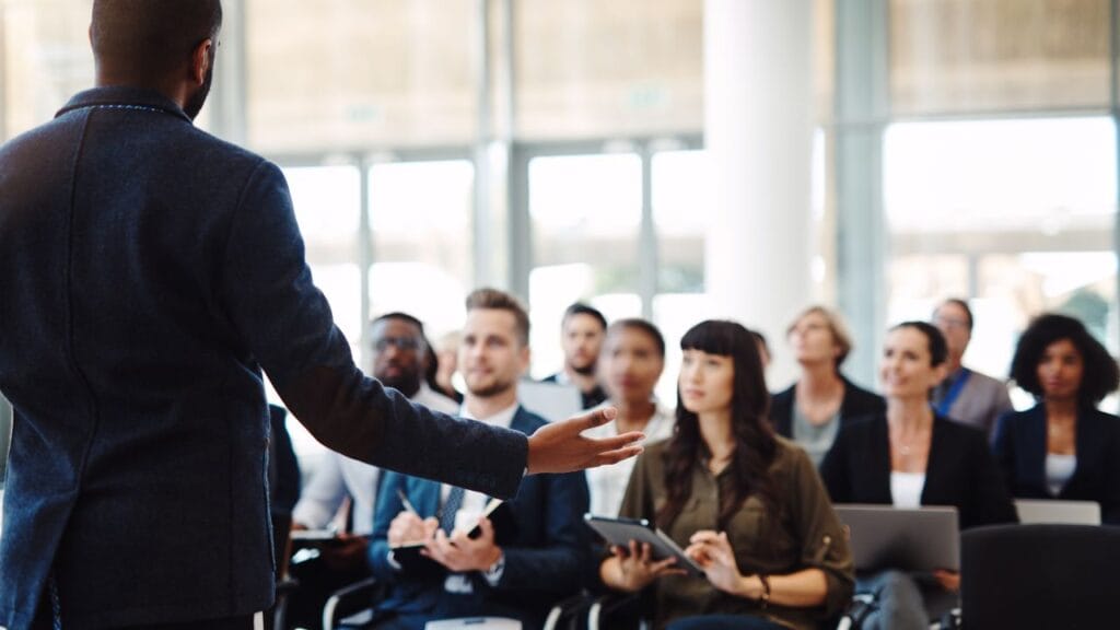 Business professional delivering a presentation to an engaged audience in a modern conference room with natural lighting. Attendees are listening attentively, taking notes on tablets and laptops.
