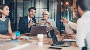 A group of five business professionals are seated around a light brown wooden conference table in a modern office setting. A woman with long brown hair sits to the left, looking towards the center of the table. A man with short dark hair, wearing a suit and tie, sits next to her, smiling and gesturing with his hands. An older woman with short gray hair and glasses, wearing a light-colored blouse and an orange scarf, sits across from them, also smiling and engaged in conversation. Another man with glasses and a beard is partially visible to the right, along with a fifth person whose back is to the viewer. Several laptops, coffee cups, and documents are scattered on the table. The background features a blurred office environment with glass walls and reflections.