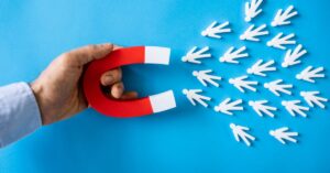 A hand holds a red horseshoe magnet against a vibrant blue background, pulling numerous small white paper cutout figures of people toward it. The figures, arranged in a scattered yet directional pattern, visually demonstrate the concept of attraction or influence. The stark contrast between the bold red magnet, pristine white figures, and cool blue backdrop emphasizes themes of leadership, connection, or strategic pull—commonly used to illustrate ideas like networking, community building, or the power of compelling vision. The simplicity of the composition ensures clarity while inviting interpretation of its metaphorical intent.