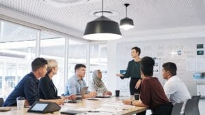 A diverse group of professionals engages in a collaborative meeting within a bright, modern office space, emphasizing why dynamic empathetic listening is critical to fostering effective communication and strategic alignment. A leader stands at the front, addressing colleagues seated around a sleek conference table, while large windows flood the room with natural light. A whiteboard behind the presenter displays handwritten notes titled "WHERE ARE WE GOING THIS YEAR?" alongside strategic diagrams and goals. Attendees appear attentive—some taking notes, others making eye contact—highlighting how active, empathetic engagement drives team cohesion and decision-making in a leadership-driven, goal-oriented environment. Key Elements for SEO: Integrates the keyword "Why Dynamic Empathetic Listening" as a central theme. Connects empathetic communication to strategic outcomes (e.g., goal-setting, team cohesion). Maintains focus on leadership, collaboration, and professional development.
