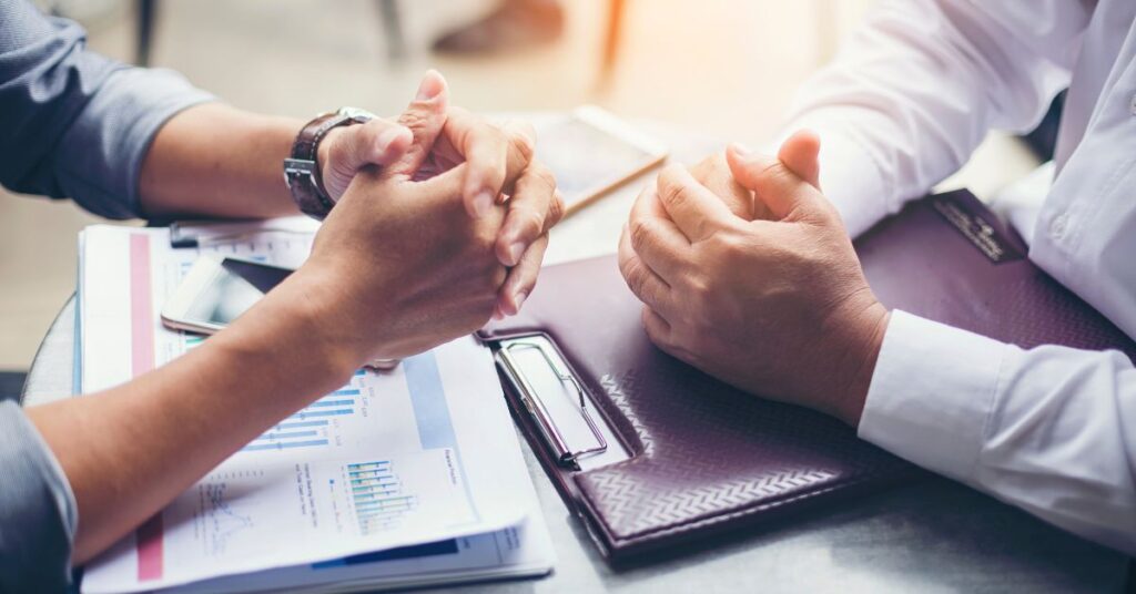 Two professionals shake hands over a cluttered conference table filled with financial reports, bar charts, and a smartphone, symbolizing a successful business agreement. Their clasped hands dominate the foreground, while one participant wears a watch and the other has a notebook nearby. The scene conveys themes of collaboration, strategic decision-making, and the culmination of negotiations within a corporate environment.