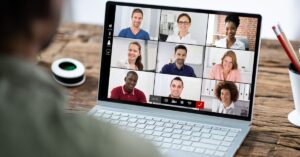 A close-up of a laptop screen showing a virtual team meeting with nine participants, including the host (visible from behind) seated at a wooden desk. The screen displays a grid of faces: a mix of men and women from diverse backgrounds, all smiling or engaged in discussion. The host wears a light gray shirt; others are dressed casually in blues, whites, and pinks. On the desk, a white mug, red pencils in a holder, and a coiled cable are visible. The laptop interface includes a timer (05:23), video controls, and participant icons, emphasizing a collaborative, remote work environment.