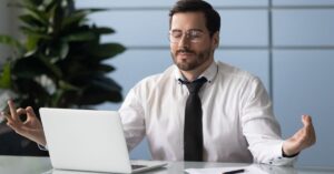 A professionally dressed man with glasses and a beard sits at a sleek desk in an office, practicing mindfulness. His eyes are closed, and his hands form meditative gestures (OK sign and open palm) while seated beside an open laptop, papers, and a pen. The background features a vibrant green potted plant and light blue tiled walls, creating a calm, modern workspace atmosphere. The image conveys the integration of relaxation techniques into daily professional life.