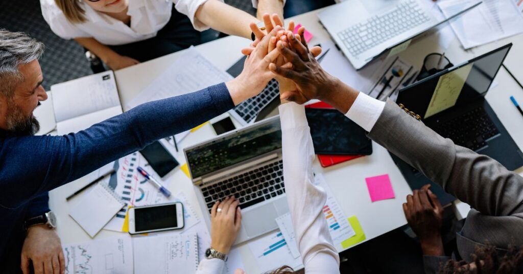 A diverse team of five professionals celebrates a collaborative achievement by stacking their hands in a unified high-five above a cluttered workspace. The overhead view reveals a dynamic meeting environment with open laptops, scattered documents, sticky notes, and mobile devices on the table. Team members wear varied business attire, and their focused yet joyful expressions emphasize camaraderie and shared success. The composition highlights teamwork, innovation, and the culmination of collective effort in a professional setting.