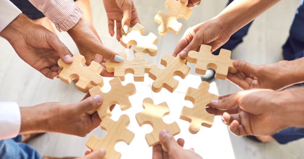 Group of individuals collaborating to assemble a wooden puzzle. Multiple hands hold interlocking pieces, symbolizing teamwork and problem-solving. The diverse participants, dressed in varied attire, contribute their unique pieces to form a cohesive whole, emphasizing unity and shared objectives." This description highlights the collaborative action, visual metaphor (puzzle pieces), and thematic elements (teamwork, unity) while incorporating SEO keywords like "teamwork" and "problem-solving.