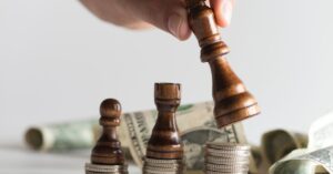 Hand holding a wooden chess rook above a strategic arrangement of chess pieces positioned on stacks of silver coins, with crumpled dollar bills in the background. The scene symbolizes financial strategy, investment decisions, and the intersection of wealth management with tactical planning, emphasizing the importance of foresight and resource allocation in achieving economic goals.