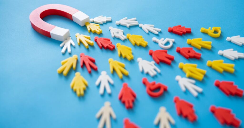 A vibrant illustration featuring a red-and-white horseshoe magnet pulling in colorful human-shaped figures on a bright blue background. The magnet attracts white, yellow, and some red figures, while others remain scattered or repelled, symbolizing selective engagement or influence. The scene emphasizes themes of attraction, diversity, and strategic targeting, with the contrasting colors highlighting varied responses to a central force—ideal for concepts like customer acquisition, team dynamics, or inclusive leadership.