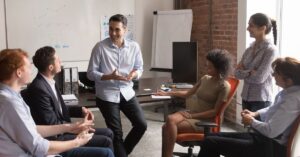 Group of six professionals engaged in a collaborative discussion around a conference table in a modern office. The central figure, a man in a light blue shirt, gestures animatedly while speaking, surrounded by attentive colleagues who nod, smile, and interact. The room features a whiteboard with charts, a flip chart, and large windows overlooking a cityscape, emphasizing a dynamic and inclusive work environment. The scene conveys themes of teamwork, strategic planning, and open communication, highlighting diverse perspectives in business decision-making.