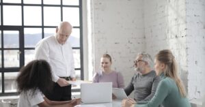 Group of five professionals collaborating in a modern office with large windows and exposed white brick walls. A bald man in a white shirt and black pants stands at the head of the table, holding documents and addressing the team. Four seated colleagues—two men and two women—engage attentively; one takes notes, another reviews a laptop, and two converse. The bright, airy space emphasizes transparency and teamwork, with sunlight streaming through the windows, highlighting a focused yet dynamic work environment centered on strategic discussion and problem-solving.