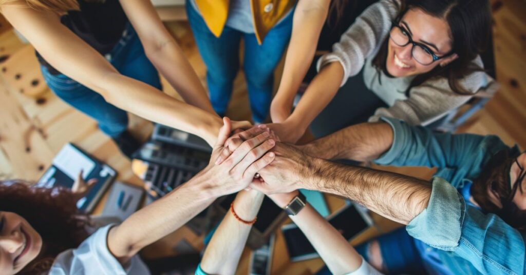 Overhead view of six professionals from diverse backgrounds stacking their hands together in a unified circle, symbolizing teamwork and collaboration. Smiling faces and engaged body language convey camaraderie and shared purpose. The scene takes place in a modern office setting with scattered laptops, notebooks, and digital devices, emphasizing a dynamic and inclusive work environment focused on collective achievement.