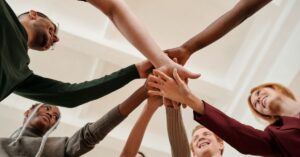 Diverse team of five professionals stacking hands in a unified pyramid formation. Participants, representing varied ethnicities and dressed in business casual attire (green long-sleeve, gray hoodie, brown sweater, maroon blazer, beige shirt), extend their arms upward toward a central focal point. Smiling faces and engaged body language convey collaboration and shared purpose. The setting features minimalist white walls and soft overhead lighting, emphasizing the symbolic gesture of solidarity and collective effort.