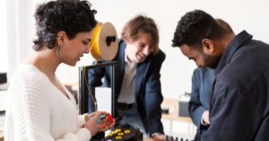 Team of professionals collaborating on a 3D printing project in a modern office. A woman in a white sweater holds a blue prototype component, while a man in a navy blazer leans over the printer, observing its operation. Another colleague adjusts settings on the machine, which sits on a wooden desk surrounded by scattered tools and materials. Large windows allow natural light to fill the room, emphasizing a creative and innovative workspace. The scene highlights hands-on problem-solving, technological experimentation, and interdisciplinary teamwork in a dynamic professional environment.