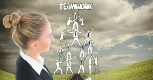 A young businesswoman in a black blazer and white blouse stands on a grassy hill, gazing thoughtfully at a towering human pyramid formed by silhouetted figures in suits. The pyramid, labeled 'TEAMWORK' at its peak, represents collaborative effort and hierarchical support. The scene is set against a dramatic sky with swirling clouds, a winding road leading toward the structure, and scattered sunlight breaking through. The imagery emphasizes themes of unity, strategic leadership, and the collective strength required to achieve shared goals, with the woman’s contemplative stance suggesting reflection on the power of teamwork.