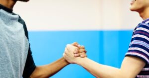Two individuals performing a fist bump in a vibrant blue setting. The person on the left wears a gray and black t-shirt, while the other sports a colorful striped shirt. Their clasped fists symbolize collaboration, agreement, or celebration, conveying enthusiasm and camaraderie against a solid blue backdrop.