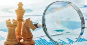 A trio of light wooden chess pieces—a king, rook, and knight—stand beside a magnifying glass on a backdrop of global maps and statistical charts, symbolizing the fusion of strategic foresight and analytical rigor in navigating complex business landscapes.