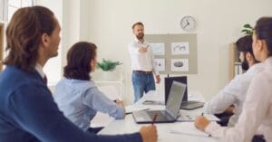 A dynamic office scene captures a collaborative meeting in progress. A man with short brown hair and a neatly trimmed beard stands confidently at the front, dressed in a crisp white button-down shirt and casual blue jeans. He holds a pen in his right hand, gesturing toward a whiteboard filled with colorful charts, graphs, and diagrams, including a prominent bar graph and pie chart. Seated around a sleek white table are four engaged colleagues: two men and two women, all focused intently on the presentation. One woman in a light blue blouse takes notes, while others lean forward, some with laptops open, indicating an interactive discussion. The room’s modern aesthetic includes a large window with blinds partially drawn, a potted plant adding a touch of greenery, and a wall clock showing 10:10 above the whiteboard. The atmosphere exudes professionalism and teamwork, emphasizing strategic planning or data analysis in a bright, airy workspace.