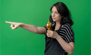 A woman with short dark hair is standing against a solid green background. She is wearing a black and white striped shirt and holding a golden trophy in her left hand, which she cradles close to her chest. With her right arm extended, she points forward with her index finger, suggesting determination or direction. Her expression appears focused and confident, conveying a sense of achievement and motivation.