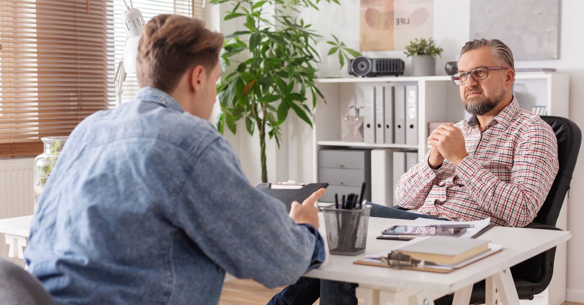 A professional consultation takes place in a well-lit office space. Two men sit across from each other at a white desk: Left : A younger man in a light blue denim shirt and dark jeans leans forward, gesturing with his right hand as he speaks. Right : An older man with a gray beard and glasses, wearing a red-and-white checkered shirt, sits back in his chair with clasped hands, listening intently. The desk holds a laptop, stacked books, a clear pencil holder, and scattered papers. Behind them, a tall green potted plant adds a touch of nature, while a white shelving unit displays organized binders, a small projector, and decorative items. Blinds cover a nearby window, allowing soft natural light to fill the room. The scene conveys a focused, collaborative atmosphere, likely centered on career guidance, strategy discussion, or personal development.