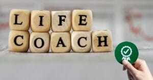 Eight light wooden blocks spell 'LIFE COACH' in two stacked tiers on a reflective surface, while a hand gently holds a green circular badge featuring a checkmark ribbon, symbolizing certified expertise. The softly blurred newspaper backdrop underscores the depth of knowledge inherent in life coaching.