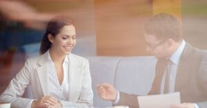 A professional meeting unfolds in a warmly lit café, where a smiling woman in a crisp white blazer engages in conversation with a man in a beige suit who reviews documents. Their relaxed postures and the ambient glow of the setting underscore a collaborative exchange in a comfortable, modern workspace.