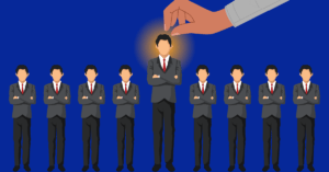 A stylized illustration features nine identical figures in gray suits with red ties, standing in a straight line against a deep blue background. The central figure is slightly taller and radiates a warm golden glow around his head, drawing attention as the focal point. Above him, a large, disembodied hand descends from the upper right corner, its index finger pointing directly at his illuminated head. The symmetry of the composition emphasizes themes of selection, leadership, or singularity—highlighting how one individual stands out amidst uniformity, possibly symbolizing recognition, potential, or distinction in a collective context.