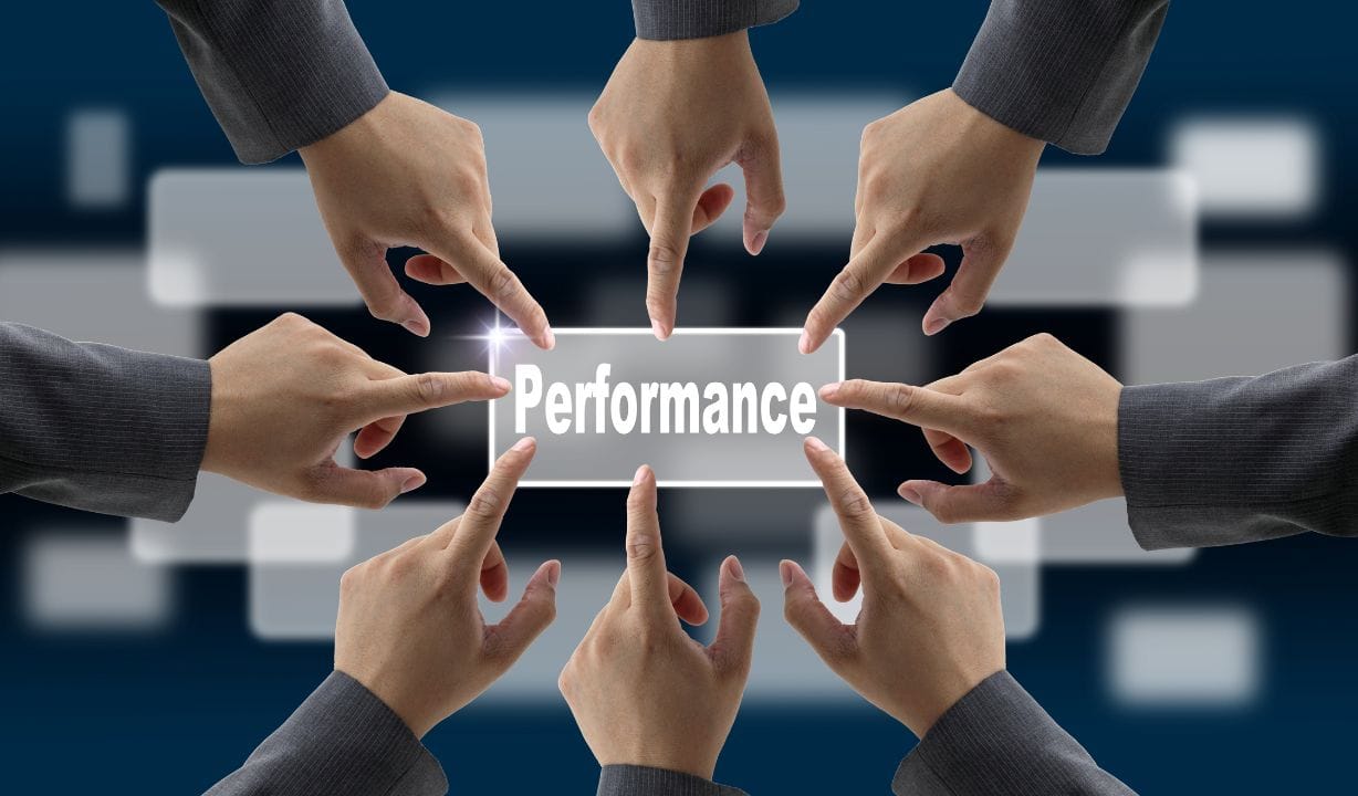 An illustration showing multiple hands pointing toward a glowing, transparent rectangular sign with the word 'Performance' written on it. The hands are reaching from different directions, symbolizing collaboration and collective focus on achieving performance goals. The background is dark and blurred, featuring abstract geometric shapes that suggest a professional or corporate setting. The overall design emphasizes teamwork and shared responsibility in driving performance.