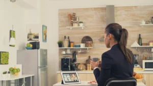 A woman is seated at a kitchen table, participating in a video conference on a tablet. She is wearing a dark blazer and holding a mug, suggesting a professional yet relaxed home environment. The tablet displays a grid of faces, indicating a virtual meeting. The kitchen setting includes a refrigerator, plants, and fruit, portraying a home office setup.