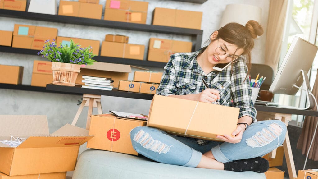 A young woman, who appears to be of East Asian descent, is sitting cross-legged on a light gray cushion in a room that seems to be a home office or a small e-commerce business setup. She is wearing round glasses, a blue and green plaid long-sleeved shirt, and ripped light blue jeans. Her hair is tied up in a bun. She is holding a brown cardboard box in her lap with a white string tied around it. In her right hand, she holds a pen and appears to be writing on a clipboard that rests on another cardboard box. Her left hand holds a smartphone to her ear, indicating she is on a call. She is smiling and looking down at what she is writing. The background shows a room with light gray walls and dark gray shelves stacked with numerous brown cardboard boxes of various sizes. There is a potted plant with purple flowers on a stand to her left and a computer monitor and other office supplies on a desk to her right. Open cardboard boxes are scattered on the floor around her. Natural light is coming from a window in the background. The scene suggests she is managing orders or inventory for her online business from home.