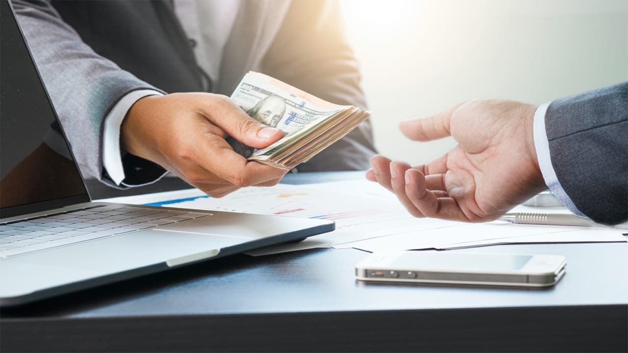 A businessman in a gray suit hands a stack of hundred-dollar bills to another businessman across a sleek black desk, symbolizing a financial transaction. The scene includes a laptop, financial documents, and a smartphone, emphasizing a professional setting for monetary exchange. Bright backlighting highlights the interaction, underscoring the transparency and formality of the deal.