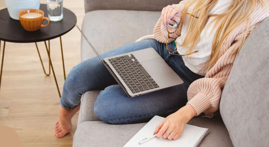 A person is comfortably seated on a gray couch, working or studying with a laptop placed on their lap. They are wearing a cozy pink sweater and blue jeans, with bare feet resting on the couch. The individual is holding a pair of glasses in one hand while the other hand rests on an open notebook with a pen lying on it, suggesting they are taking notes or preparing to write. In the background, there is a small round table with a cup of coffee and a glass of water, adding to the relaxed and productive atmosphere of the setting.