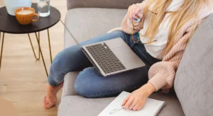 A person is comfortably seated on a gray couch, working or studying with a laptop placed on their lap. They are wearing a cozy pink sweater and blue jeans, with bare feet resting on the couch. The individual is holding a pair of glasses in one hand while the other hand rests on an open notebook with a pen lying on it, suggesting they are taking notes or preparing to write. In the background, there is a small round table with a cup of coffee and a glass of water, adding to the relaxed and productive atmosphere of the setting.