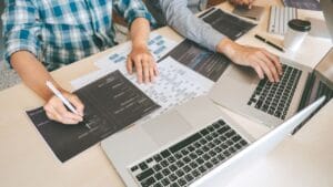 Two individuals are engaged in a collaborative coding or design session at a desk. One person, wearing a blue plaid shirt, is holding a pen and pointing to a printed document with code or design elements on it. The other individual, dressed in a light gray shirt, is actively typing on a laptop keyboard. The workspace includes multiple laptops displaying code, scattered papers, and a coffee cup, suggesting a focused and productive environment. The setting appears to be a modern office or workspace, emphasizing teamwork and technical work.