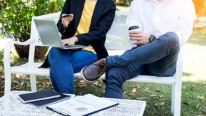 Two individuals are seated on a white outdoor bench in a park-like setting, engaged in a professional conversation. One person is using a laptop and holding a pen, while the other holds a coffee cup. A small table in front of them is adorned with notebooks, papers, and documents, suggesting a collaborative work session. The background features greenery and sunlight, creating a relaxed yet productive atmosphere.