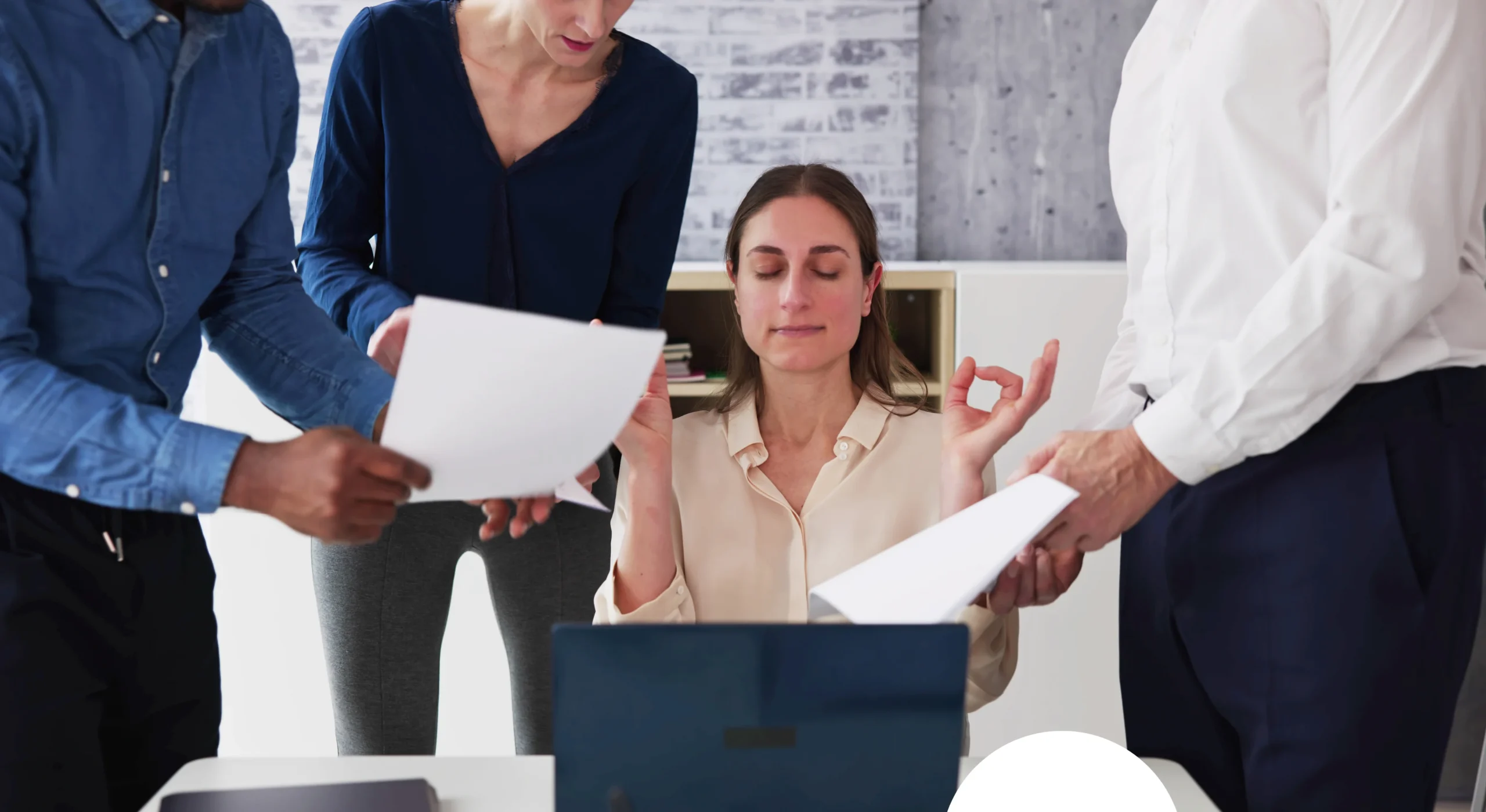 A woman is seated at a desk in an office setting, appearing calm and focused as she practices meditation or deep breathing. She has her eyes closed and hands positioned in a meditative gesture, suggesting mindfulness or stress reduction techniques. Surrounding her are three colleagues who are engaged in work activities: one person on the left is holding a document, another in the middle is also holding papers, and the third on the right is gesturing while holding additional documents. The background includes a whiteboard with notes and diagrams, indicating a collaborative workspace. The scene highlights the integration of mindfulness practices into a busy professional environment.