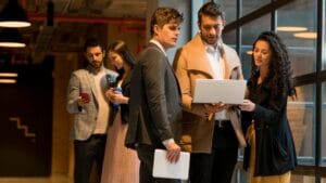 A group of professionals in a modern office setting, collaborating and working together. Two individuals are focused on a laptop screen, while others hold devices or engage in conversation. The environment features exposed ceilings, industrial lighting, and large windows with an urban view.