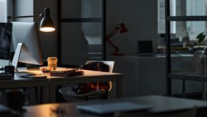 Workplace communication issues: Lit office desk at night. Computer, lamp, city view.