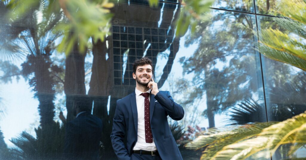 Man in a suit on a phone outside a glass building with palms.