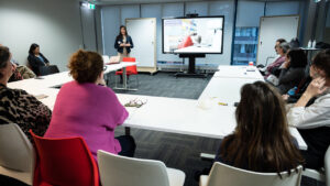 A group meeting with a presenter discussing "How Toronto Coaches Use NLP to Unlock Success" on a screen, attended by people around a conference table.