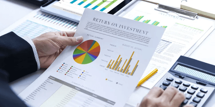 Business person using a calculator while reviewing charts and graphs to track ROI on investment reports.