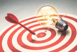 A dart hits a red and white target near the bullseye, next to a glowing lightbulb, questioning Traditional Goal-Setting methods.